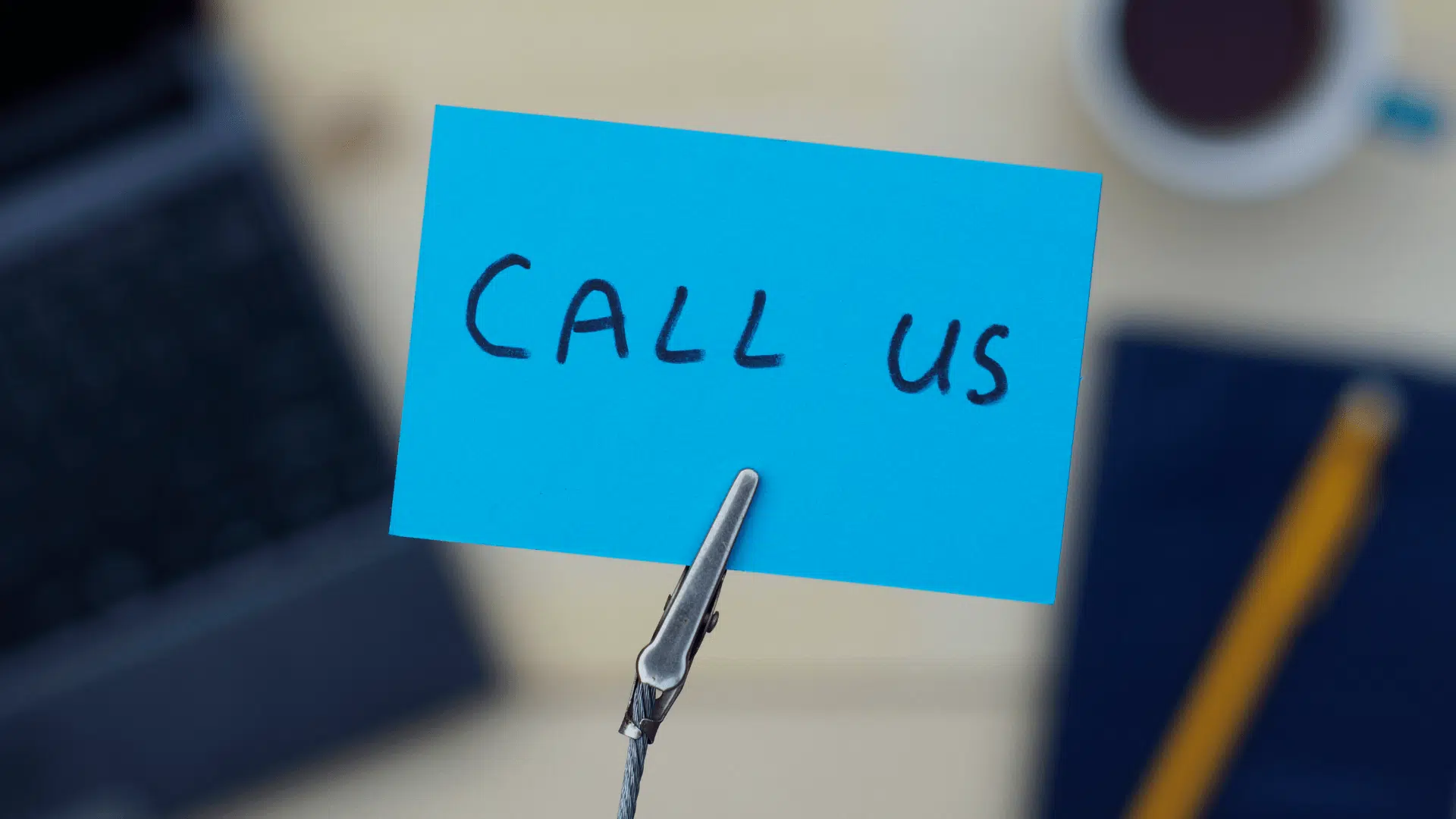 Blue sticky note with "call us" written on it, clipped to a stand with blurred laptop and coffee cup in the background.