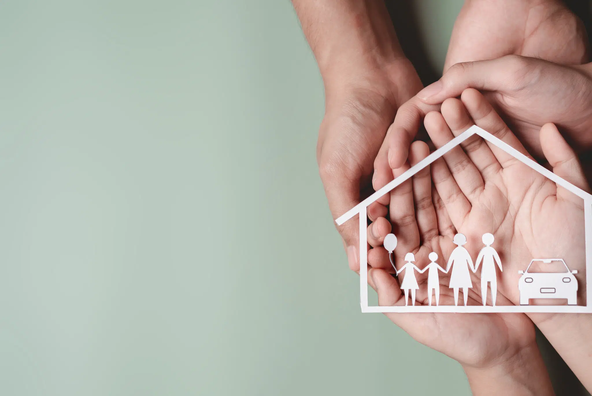 Hands holding a paper cutout of a house and family over a soft green background, symbolizing home safety and insurance.