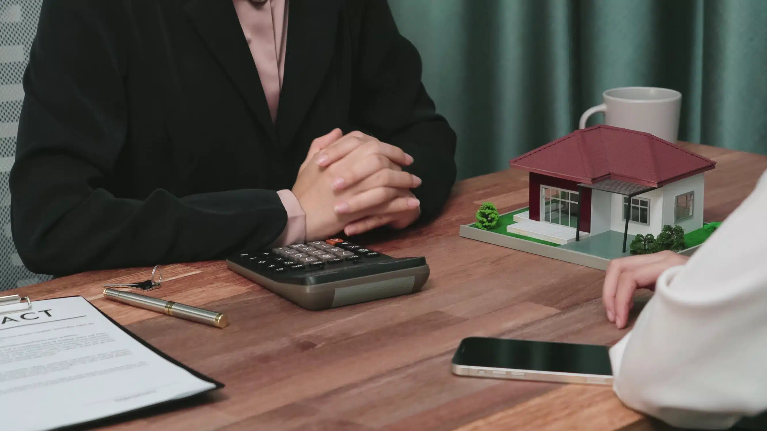 Two people at a table discussing a real estate transaction, with a contract, calculator, and model house present.